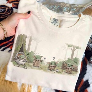 May include: Cream-colored sweatshirt featuring a forest scene with cartoon characters. The graphic depicts several characters in brown robes, with one in a pod. The word "COOl" is written on a log. The Comfort Colors tag is visible.