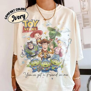 May include: Ivory-colored t-shirt featuring the Toy Story logo and characters. The design includes Woody, Buzz Lightyear, Jessie, Rex, Bo Peep, and the Little Green Men. The phrase "You've got a Friend in me" is also printed on the shirt.