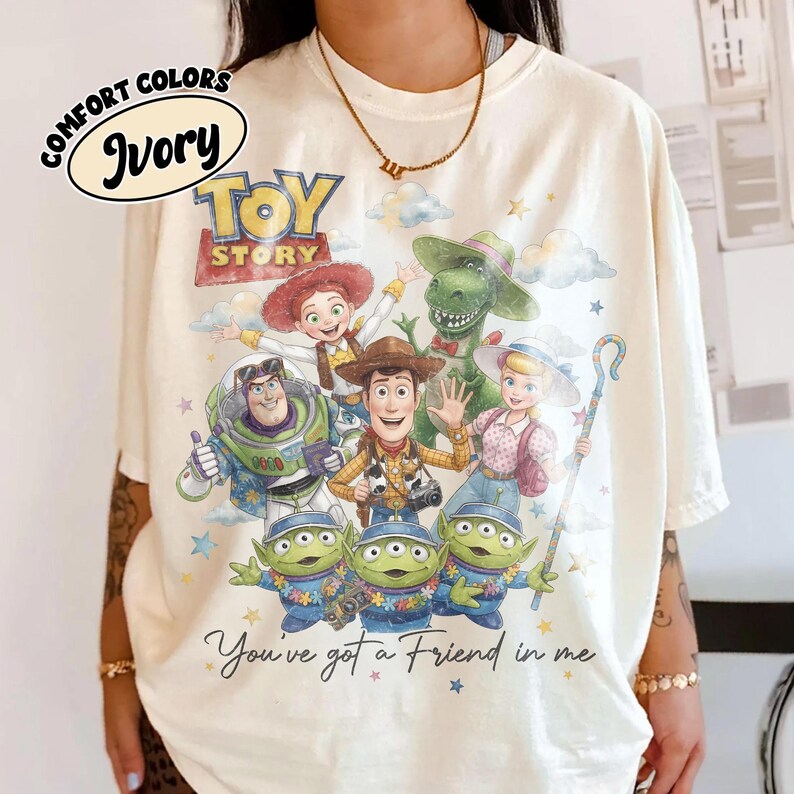 May include: Ivory-colored t-shirt featuring the Toy Story logo and characters. The design includes Woody, Buzz Lightyear, Jessie, Rex, Bo Peep, and the Little Green Men. The phrase "You've got a Friend in me" is also printed on the shirt.