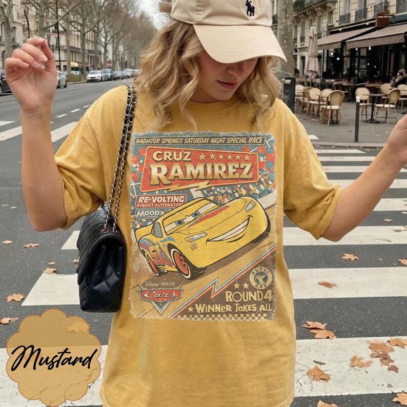 May include: Mustard-colored t-shirt featuring a graphic of Cruz Ramirez from the Disney Pixar Cars movie. The design includes text that reads "RADIATOR SPRINGS SATURDAY NIGHT SPECIAL RACE" and "ROUND 4 WINNER TAKES ALL".