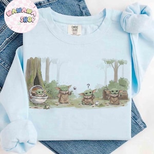 May include: Light blue Comfort Colors sweatshirt featuring a graphic of a character from a popular science fiction series. The design shows the character in various poses in a forest setting. The sweatshirt has the shop's logo in the upper left corner.