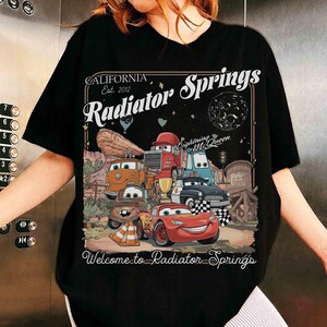Op de afbeelding: Zwart t-shirt met een kleurrijke afbeelding van personages uit de film Cars, waaronder Lightning McQueen. Het ontwerp bevat de tekst "CALIFORNIA Est. 2012 Radiator Springs" en "Welcome to Radiator Springs".