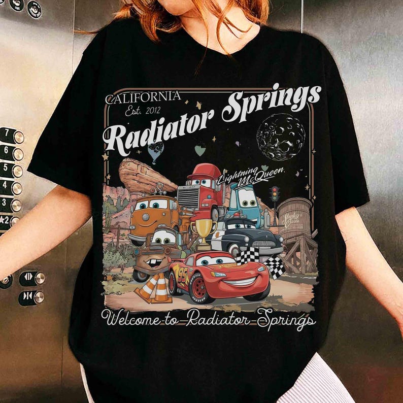 Op de afbeelding: Zwart t-shirt met een kleurrijke afbeelding van personages uit de film Cars, waaronder Lightning McQueen. Het ontwerp bevat de tekst "CALIFORNIA Est. 2012 Radiator Springs" en "Welcome to Radiator Springs".