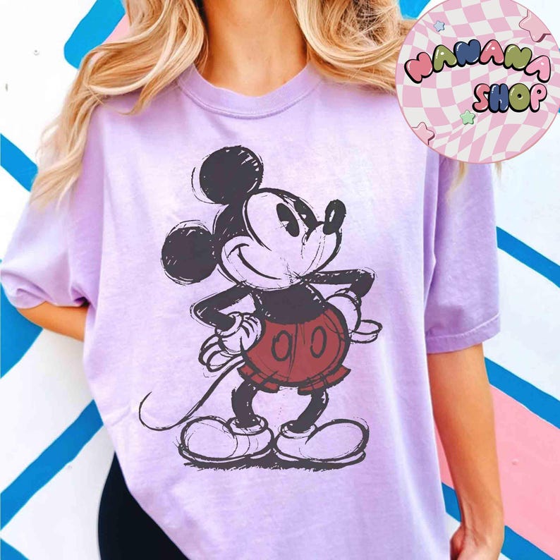 K&ouml;nnte beinhalten: Ein helllila T-Shirt mit einem skizzierten Mickey Mouse-Design in Schwarz, Rot und Wei&szlig;. Das Oberteil hat kurze &Auml;rmel und eine lockere Passform. Der Text "WANANA SHOP" befindet sich in einem Kreis oben rechts.