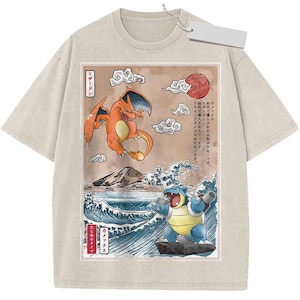 May include: A beige short-sleeve t-shirt featuring a graphic print of two creatures, one orange and one blue, set against a landscape with a mountain, ocean waves, and clouds. Japanese text is printed on the left and right sides of the graphic.
