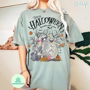 May include: Sage green t-shirt featuring a graphic from "The Nightmare Before Christmas." The design includes Jack Skellington, Sally, Oogie Boogie, and other characters, with the text "This is Halloween." Pumpkins and bats are also included.
