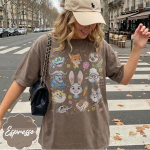 Pu&ograve; includere: T-shirt color espresso con illustrazioni di personaggi dei cartoni animati. La maglietta mostra vari personaggi di animali con testo scritto a mano. La maglietta &egrave; indossata con un berretto beige e una borsa nera.