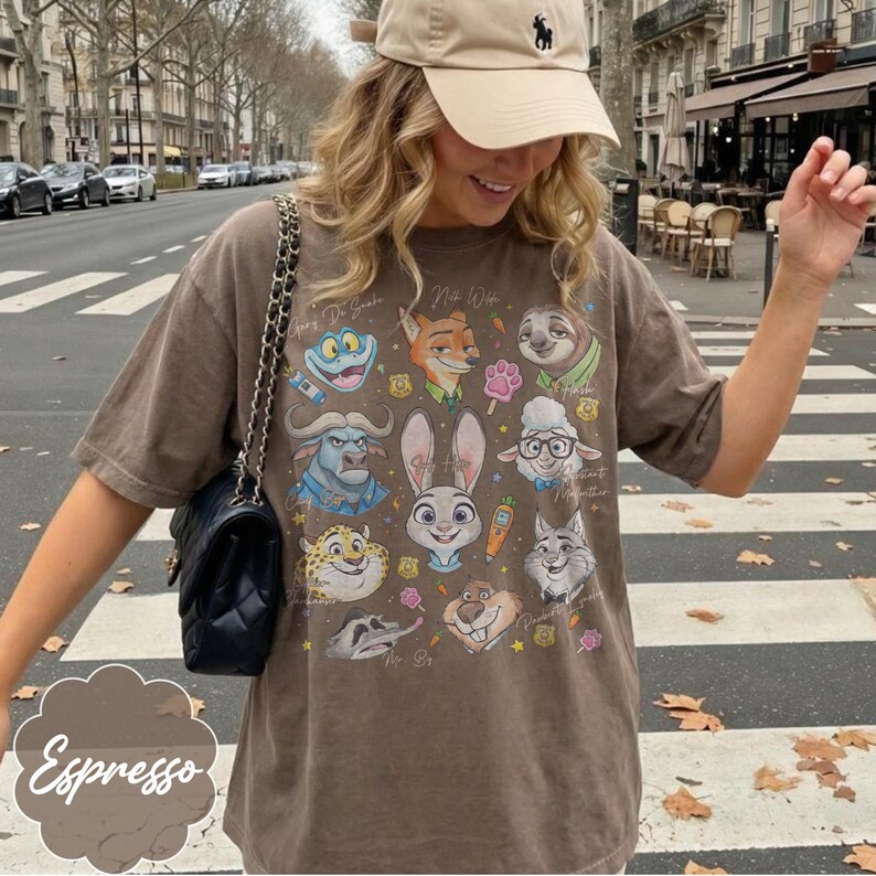 Pu&ograve; includere: T-shirt color espresso con illustrazioni di personaggi dei cartoni animati. La maglietta mostra vari personaggi di animali con testo scritto a mano. La maglietta &egrave; indossata con un berretto beige e una borsa nera.