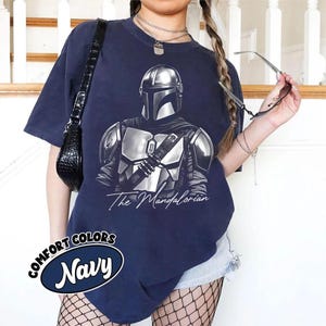 K&ouml;nnte beinhalten: Marineblaues T-Shirt mit einer detaillierten silbernen Illustration von The Mandalorian. Der Text "The Mandalorian" steht darunter in einer kursiven Schriftart. Das Shirt hat auch ein marineblaues Oval mit den Worten "Comfort Colors" und "Navy".