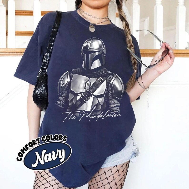 K&ouml;nnte beinhalten: Marineblaues T-Shirt mit einer detaillierten silbernen Illustration von The Mandalorian. Der Text "The Mandalorian" steht darunter in einer kursiven Schriftart. Das Shirt hat auch ein marineblaues Oval mit den Worten "Comfort Colors" und "Navy".