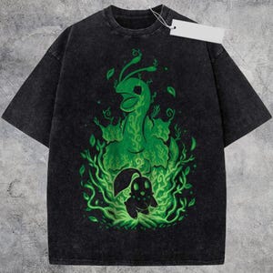 May include: A black acid-wash t-shirt featuring a central graphic of a large green creature with vine-like patterns and a smaller dark silhouette creature below, both surrounded by swirling green foliage and leaves, with a blank white price tag attached.