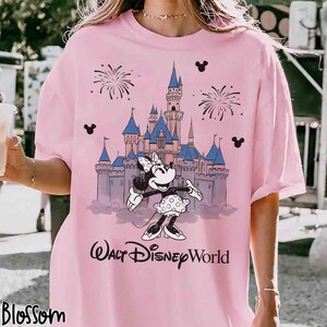 K&ouml;nnte beinhalten: Hellrosa T-Shirt mit einem Aufdruck von Minnie Mouse vor einem Schloss mit Feuerwerk und dem Text "Walt Disney World". Das Design beinhaltet Mickey Mouse Silhouetten.