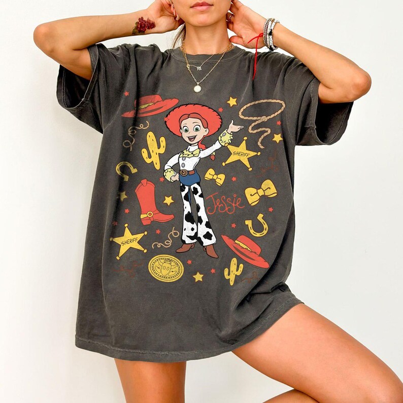 K&ouml;nnte beinhalten: Dunkelgraues T-Shirt mit einem Cartoon-Bild von Jessie aus Toy Story. Das Design umfasst einen roten Hut, Stiefel und den Namen "Jessie". Weitere Grafiken sind Sterne, ein Lasso und ein Hufeisen. Das T-Shirt ist &uuml;bergro&szlig;.