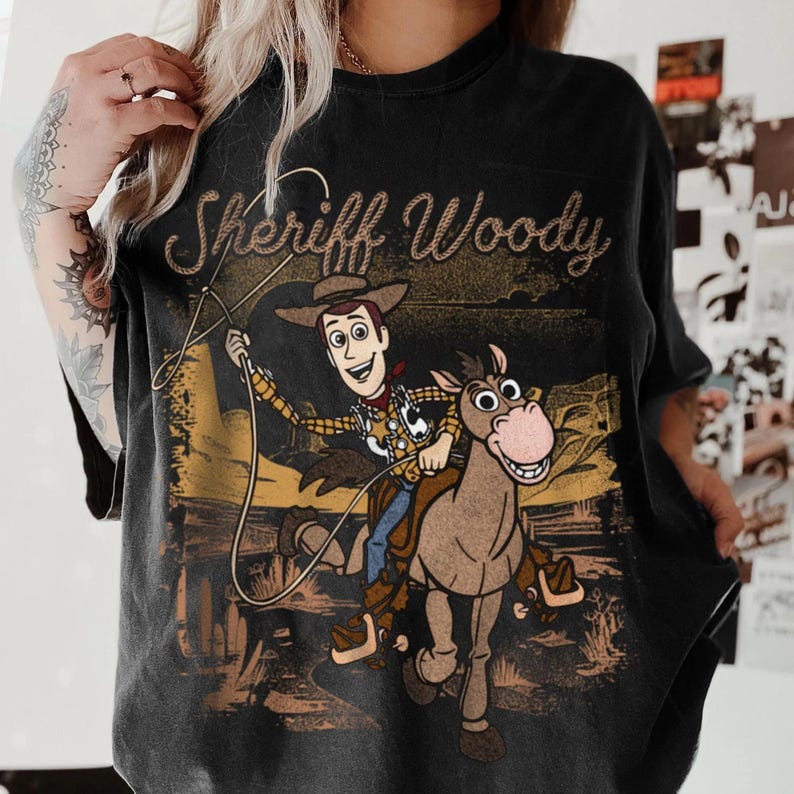 May include: Black graphic t-shirt featuring Sheriff Woody from Toy Story riding a horse. The design includes the text "Sheriff Woody" in a gold, textured font, and a desert landscape background. The shirt is a dark, faded color.