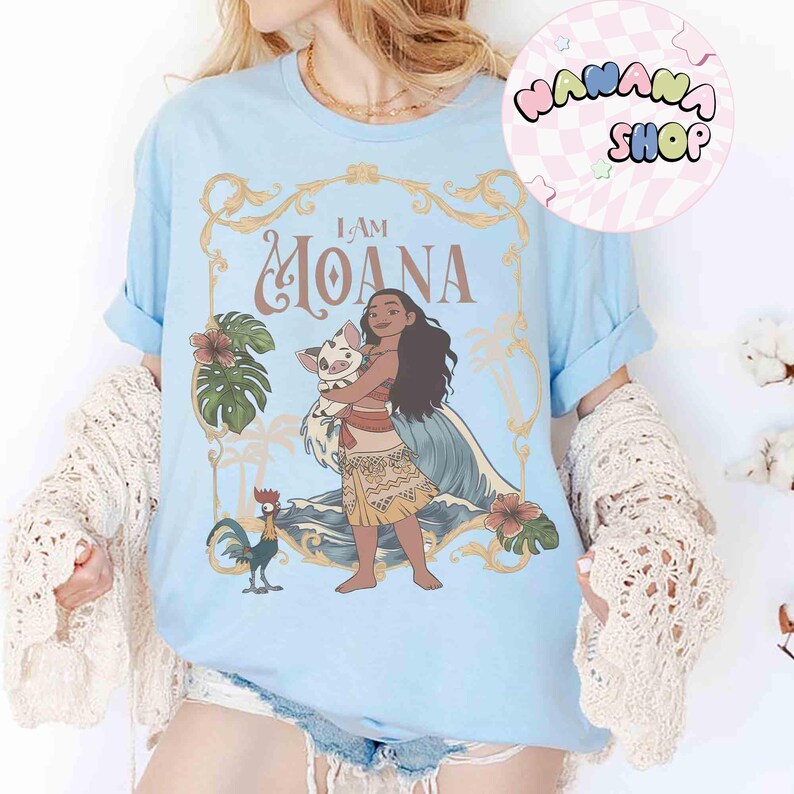 May include: Pale blue t-shirt with the words "I AM MOANA" and a graphic of Moana with a pig, a rooster, and tropical plants. The shirt has rolled sleeves and a crew neck.