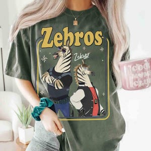 以下が含まれることがあります： 2匹のシマウマの漫画イラストが描かれたオリーブグリーンのTシャツ。「Zebros」の文字が黄色でシマウマの上に、下に「Zebros」の文字が繰り返されています。1匹のシマウマはサングラスと警察の制服を着ています。もう1匹のシマウマはレターマンジャケットを着ています。