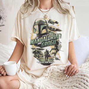 May include: Cream-colored t-shirt featuring a graphic from the Star Wars series, "The Mandalorian and Grogu." The design includes the Mandalorian's helmet, Grogu, and the phrase "THIS IS THE WAY."