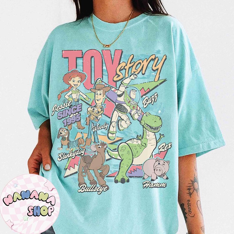 Puede incluir: Camiseta azul claro con un dise&ntilde;o de personajes de Toy Story, incluyendo a Woody y Buzz Lightyear. Las palabras "TOY STORY" est&aacute;n en rosa y rojo. Tambi&eacute;n se ve el texto "Desde 1995". La camiseta tiene un aspecto vintage.