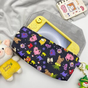 May include: A black zippered pouch featuring a colourful pattern of various cartoon creatures, including pink, purple and yellow characters. A yellow handheld gaming console is partially inside the pouch, and a green character keychain is attached.