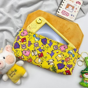 May include: A yellow Nintendo Switch Lite console is in a yellow and orange case with a cartoon character pattern. A small notepad with a bear illustration and a plush pig toy are also visible.