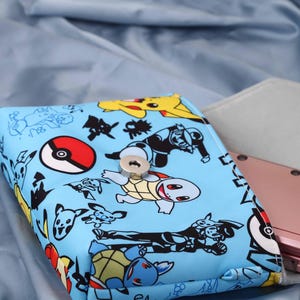 May include: A light blue case featuring various cartoon characters and a red and white design. A pink handheld gaming device is visible inside a gray case. The case has a silver clasp.