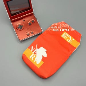 May include: A metallic red Game Boy Advance SP handheld gaming console with a flip-top screen and a matching red fabric pouch featuring a white leaf graphic, the text "NEW RV", and an orange stripe with white Japanese characters printed on it.