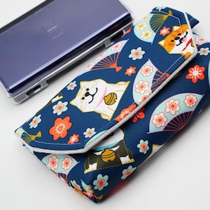 May include: A rectangular fabric pouch with a dark blue background featuring a repeating pattern of stylised dogs, fans, and orange and white flowers. The pouch has a folded flap closure with white trim and sits next to a dark purple handheld console.