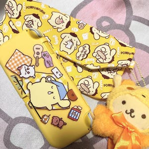May include: A yellow gaming console case and a matching yellow pouch, both featuring Pompompurin character prints and text. A small, fuzzy yellow plush toy with an orange ribbon is positioned next to the case and pouch on a patterned surface.
