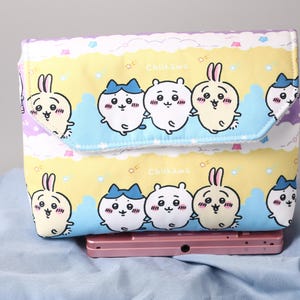 May include: A rectangular, fabric case with a flap closure, featuring a repeating pattern of cartoon characters. The design includes white, yellow, and blue colors. The case is resting on a pink electronic device, suggesting it's a protective cover.