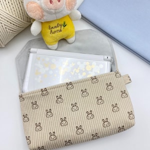Peut inclure: Une pochette en velours côtelé beige avec un motif répétitif de visages de lapins dessinés en brun. La pochette est ouverte, révélant une console de jeu portable blanche avec un motif circulaire doré et blanc à l'intérieur.