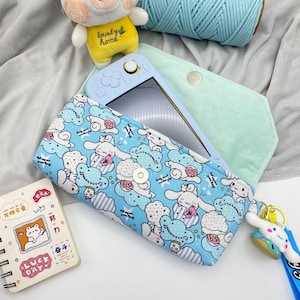 May include: A light blue fabric carrying case featuring a repeating pattern of white cartoon characters, clouds, and pink flowers. The case has a snap closure and a matching character keychain. A light blue handheld gaming console is tucked inside.