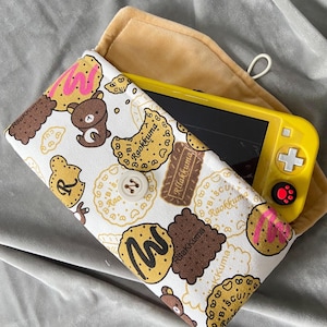 May include: A white fabric carrying case featuring a repeating pattern of brown Rilakkuma bears and various cookies with pink and brown icing. The case has a central white button closure and contains a yellow handheld gaming console with black screen.