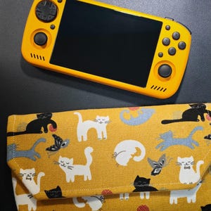 May include: A bright yellow handheld gaming console with a black screen, directional pad, analog sticks, and action buttons, positioned above a rectangular fabric pouch featuring a repeating pattern of white, black, and blue cats on a yellow background.