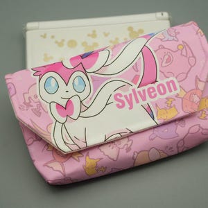 May include: A pink zipped pouch featuring a repeating pattern of various cartoon creatures and a large graphic of the character Sylveon with the text Sylveon printed on the front flap. A white handheld gaming console is visible behind the pouch.