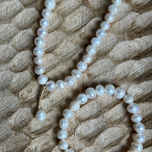 May include: A necklace and bracelet set featuring white, irregularly shaped pearls. The necklace has a gold-coloured clasp and a single pearl pendant. The jewellery rests on a textured, light brown wooden surface.
