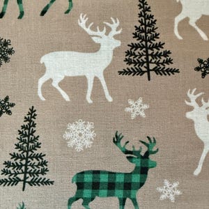 Op de afbeelding: Beige stof met een herhalend patroon van witte herten, groen en zwart geruite herten, zwarte dennenbomen en witte sneeuwvlokken. Ideaal voor feestelijke knutselprojecten.