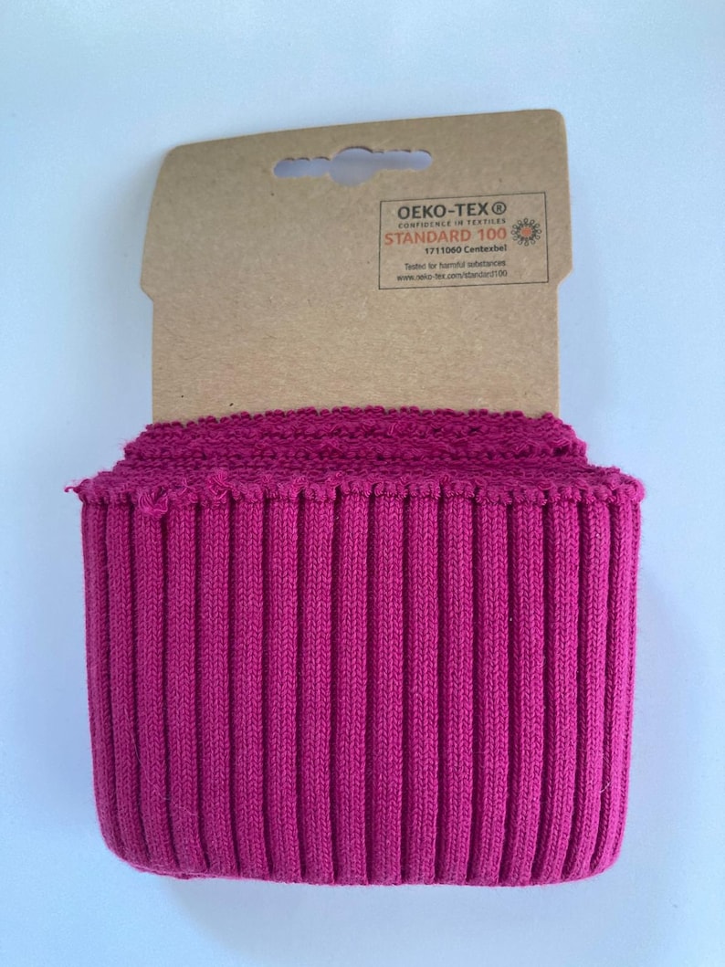 May include: A roll of vibrant fuchsia ribbed knit fabric. The fabric is tightly woven with vertical ridges, and is attached to a cardboard tag with the "OEKO-TEX&reg; STANDARD 100" label.