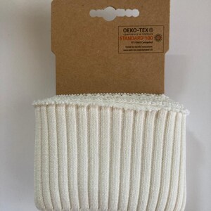 May include: Cream-colored ribbed knit fabric with a decorative edge, attached to a brown cardboard tag. The tag features the Oeko-Tex Standard 100 logo and text. The fabric is tightly knit with vertical ribs, suggesting a cuff or trim for clothing or accessories.