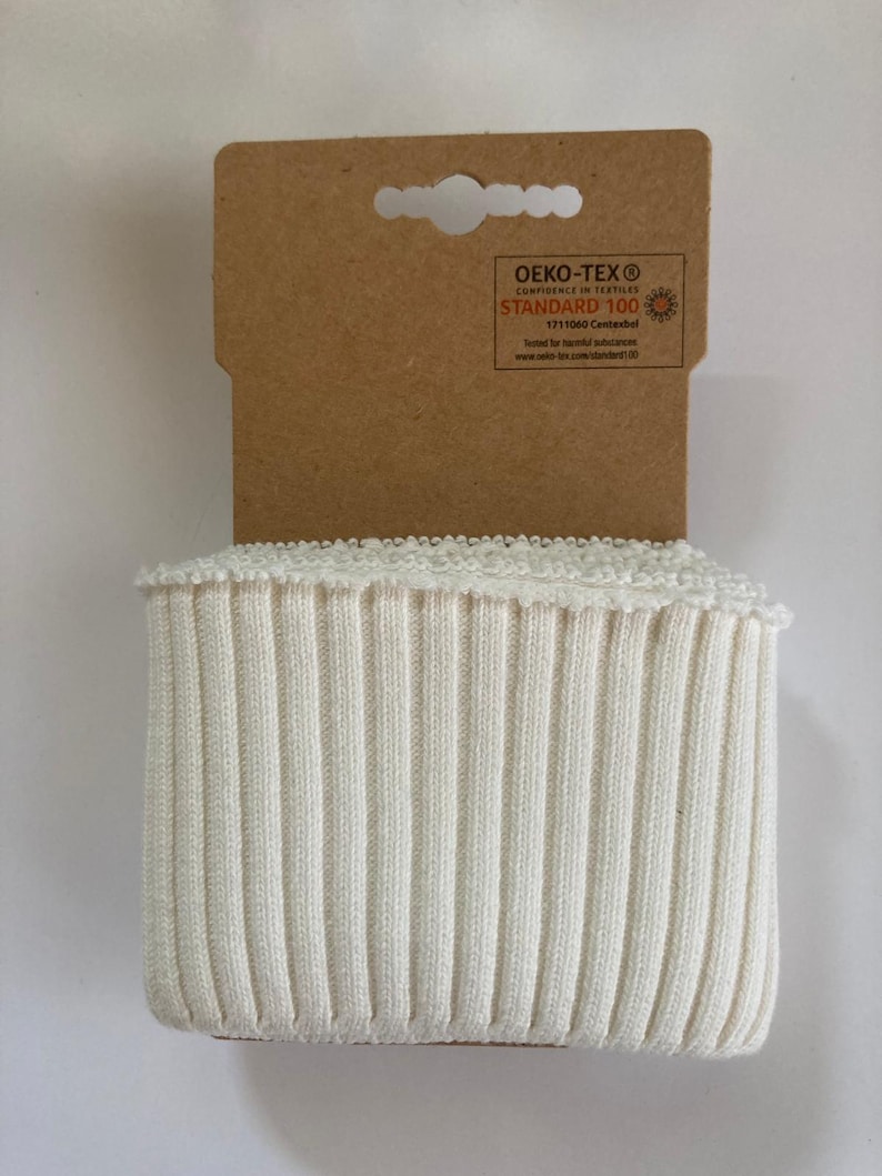 May include: Cream-colored ribbed knit fabric with a decorative edge, attached to a brown cardboard tag. The tag features the Oeko-Tex Standard 100 logo and text. The fabric is tightly knit with vertical ribs, suggesting a cuff or trim for clothing or accessories.