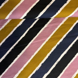 Pode incluir: Tecido com listras diagonais em amarelo mostarda, azul marinho, preto e rosa, separadas por pinceladas brancas. O tecido está dobrado, mostrando o padrão repetido.