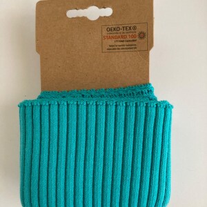 May include: A roll of turquoise blue ribbed knit fabric with a decorative edge, attached to a brown cardboard tag. The fabric is tightly rolled, showcasing its texture and color. The tag has printed text.