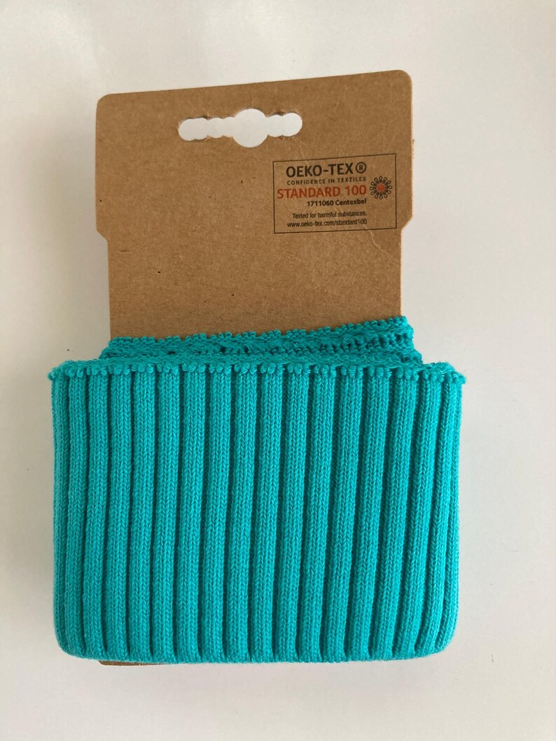 May include: A roll of turquoise blue ribbed knit fabric with a decorative edge, attached to a brown cardboard tag. The fabric is tightly rolled, showcasing its texture and color. The tag has printed text.