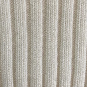 May include: Close-up of a cream-colored, ribbed knit fabric. The vertical ribs create a textured pattern, with subtle variations in the yarn's color. The fabric appears soft and likely suitable for clothing or home decor.