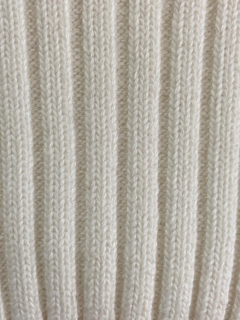 May include: Close-up of a cream-colored, ribbed knit fabric. The vertical ribs create a textured pattern, with subtle variations in the yarn's color. The fabric appears soft and likely suitable for clothing or home decor.
