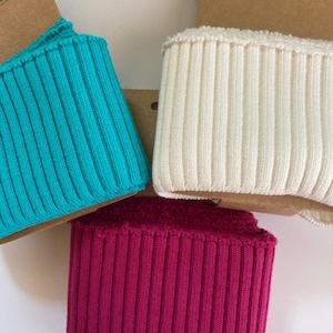 May include: Close-up of three ribbed knit fabric pieces in turquoise, off-white, and magenta. The fabric is folded over a cardboard backing. The knit has vertical lines. The Oeko-Tex Standard 100 label is visible.