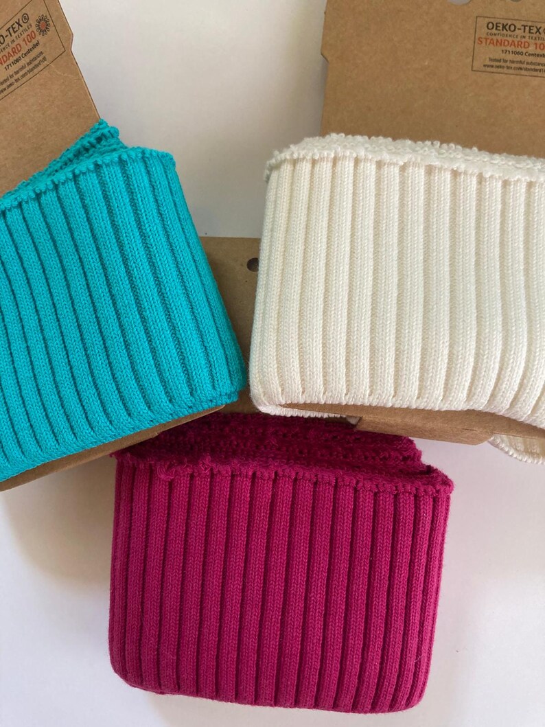 May include: Close-up of three ribbed knit fabric pieces in turquoise, off-white, and magenta. The fabric is folded over a cardboard backing. The knit has vertical lines. The Oeko-Tex Standard 100 label is visible.