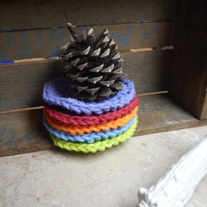 May include: A stack of six colorful crocheted coasters in shades of green, blue, orange, and purple. A brown pine cone sits on top of the coasters.