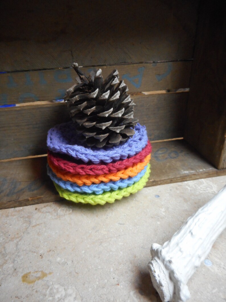 May include: A stack of six colorful crocheted coasters in shades of green, blue, orange, and purple. A brown pine cone sits on top of the coasters.