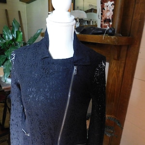 May include: A black lace jacket with an asymmetrical zipper closure and a notched collar. The jacket is displayed on a white mannequin and features long sleeves with a delicate floral pattern. The jacket has silver zippers and a tailored fit.