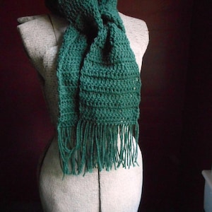 May include: A green crocheted scarf with fringe on the ends. The scarf is tied around a mannequin's neck.